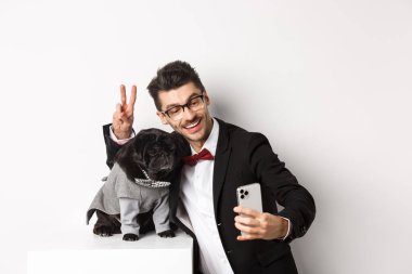 Takım elbiseli neşeli köpek sahibi köpekle yeni yılı kutluyor. Akıllı telefondan özçekim yapıyor. Kostümlü, beyaz arka planlı, sevimli siyah bir köpeğin yanında.