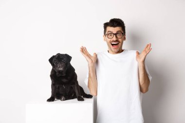 Gözlüklü neşeli genç adam, köpeğiyle ayakta, neşe içinde kameraya bakıyor, eğleniyor, harika haberler duyuyor, beyaz arka planda pug ile ayakta duruyor.