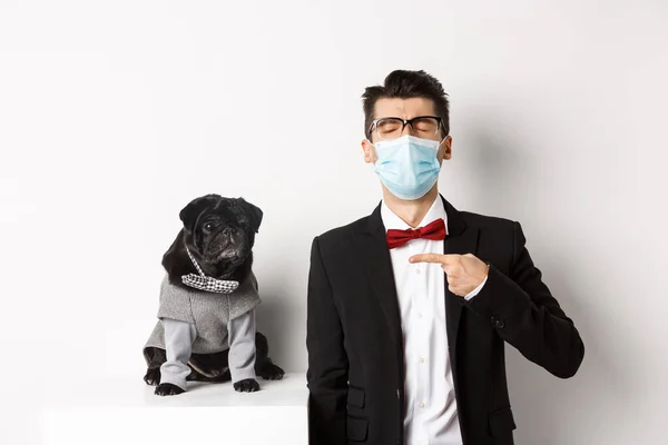 Coronavirus, pets and celebration concept. Disappointed young man in face mask and suit, pointing finger at cute black pug dog wearing party costume, standing over white background