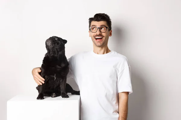 Ung mann med briller som klemmer hunden sin, huseieren og hunden som stirrer på reklametilbudet i øvre venstre hjørne, stående over hvit bakgrunn – stockfoto