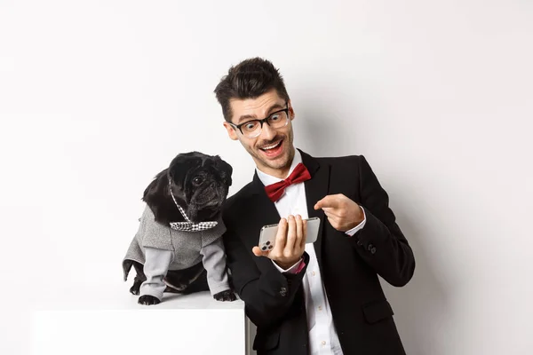 Lykkelige hundeeiere som viser noe å klappe på mobilen, mann og hund i flotte kostymer, står over hvit bakgrunn – stockfoto
