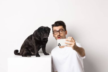 Yakışıklı bir delikanlının akıllı telefondan siyah bir köpekle selfie çekip beyaz arka planda pug pozu vermesi.