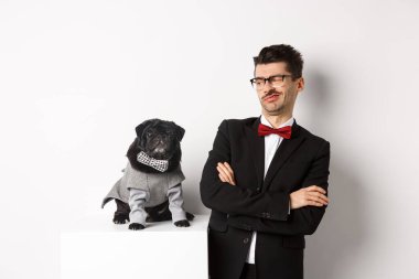 Hayvanlar, parti ve kutlama konsepti. Takım elbiseli ve gözlüklü komik bir genç adam. Kostümlü şirin köpeğe şüpheyle bakıyor. Beyaz arka planda duruyor.