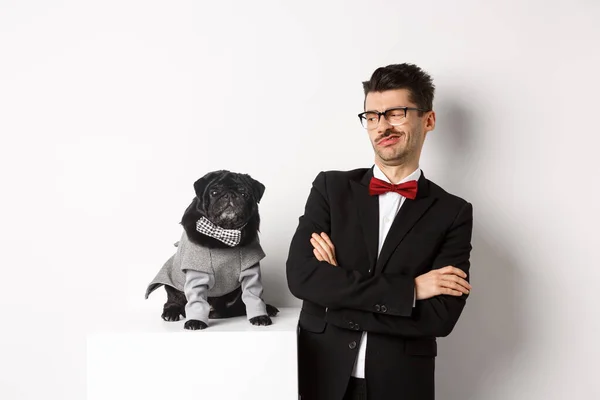 Dieren, feest en feest concept. Afbeelding van grappige jongeman in pak en bril, op zoek sceptisch op schattige pug in kostuum, staande over witte achtergrond — Stockfoto