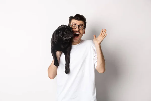 Stilig ung man i glasögon håller sin svarta mops och viftande hand, killen säger hej medan bära hund med en arm, står över vit bakgrund — Stockfoto