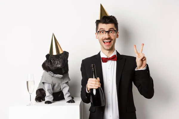 Pequeno cão preto vestindo chapéu de festa e de pé perto do homem feliz celebrando férias, proprietário mostrando sinal de paz e segurando garrafa de champanhe, fundo branco — Fotografia de Stock