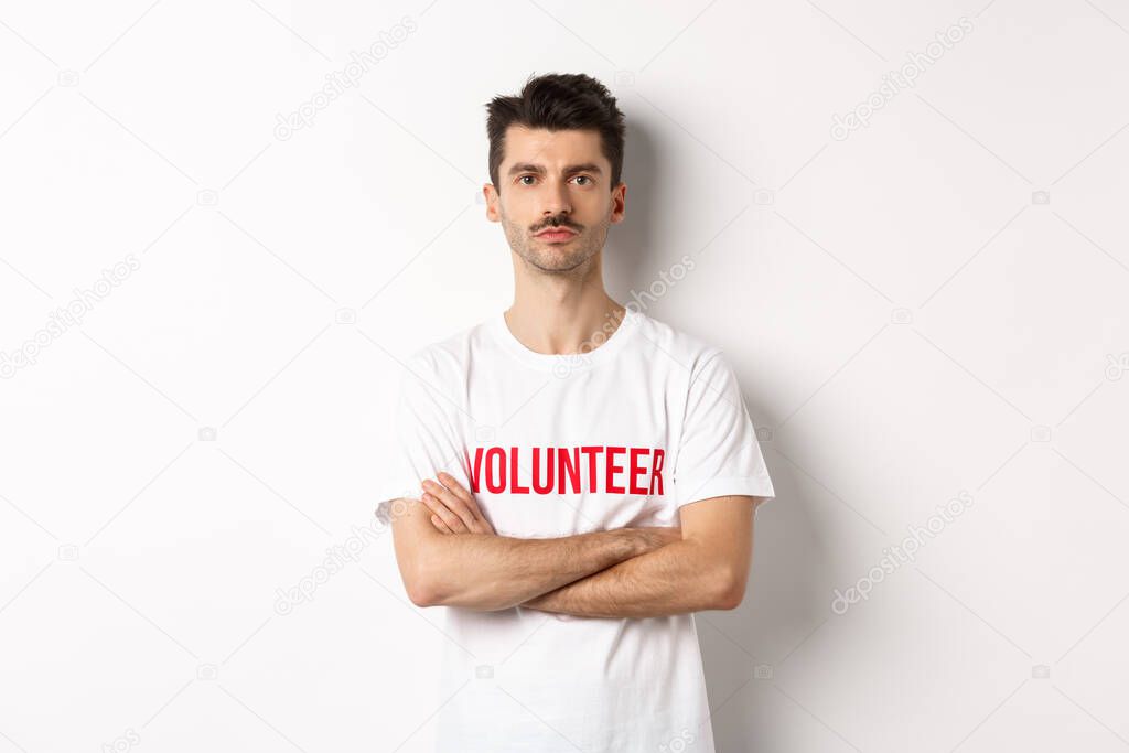 Serious young male volunteer in white t-shirt, holding arms crossed on chest, looking at camera, ready to help