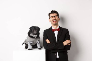 Hayvanlar, parti ve kutlama konsepti. Üzgün köpek sahibi ağlıyor, takım elbise giyiyor, kostümlü sevimli siyah bir köpeğin yanında duruyor, beyaz arka planda duruyor.