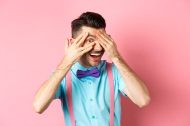 İlgi çekici papyonlu komik adam, parmaklarının arasından ilginç bir şeye bakıyor, havalı bir şeye bakıyor, pembe arka planda duruyor.