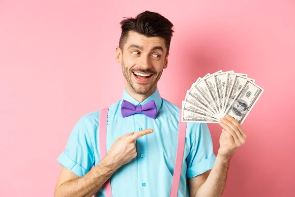 Hombre guapo rico en traje festivo apuntando al dinero, sosteniendo dólares y luciendo feliz, de pie sobre fondo rosa — Foto de Stock