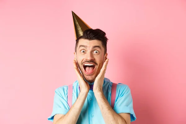 Vacaciones y concepto de celebración. Primer plano de chico divertido en sombrero de cumpleaños gritando sorprendido, gritando de alegría, recibiendo regalo impresionante, de pie feliz sobre fondo rosa — Foto de Stock