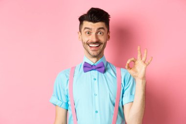 Bıyıklı yakışıklı beyaz adam gülümsüyor ve işaret veriyor, iyi bir şeyi onaylıyor, mükemmel seçimi övüyor, pembe arka planda eğleniyor.
