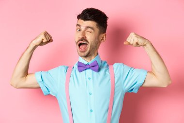 Fransız bıyıklı güçlü ve komik adam, kaslarını esnetiyor ve kameraya göz kırpıyor, güçlü yanlarını gösteriyor, pembe arka planda duruyor.