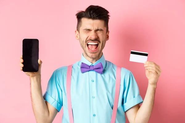 Concepto de comercio electrónico y compras. Hombre llorando mostrando la pantalla vacía del teléfono inteligente y la tarjeta de crédito de plástico, sintiéndose frustrado, de pie sobre fondo rosa — Foto de Stock