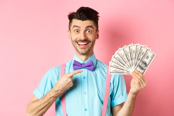 Retrato de tipo alegre con bigote y pajarita apuntando al dinero, sosteniendo dólares y sonriendo excitado, de pie sobre fondo rosa — Foto de Stock