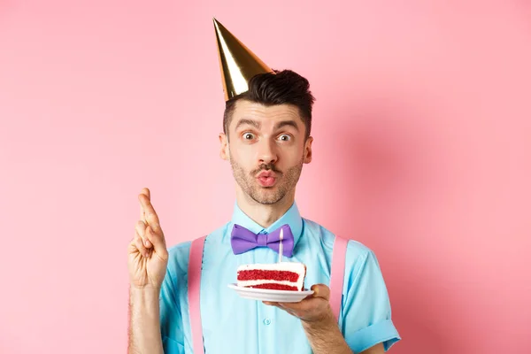 Vacaciones y concepto de celebración. Hombre lindo y divertido haciendo deseo de cumpleaños con los dedos cruzados, sosteniendo el pastel del día b y mirando soñador a la cámara, fondo rosa — Foto de Stock