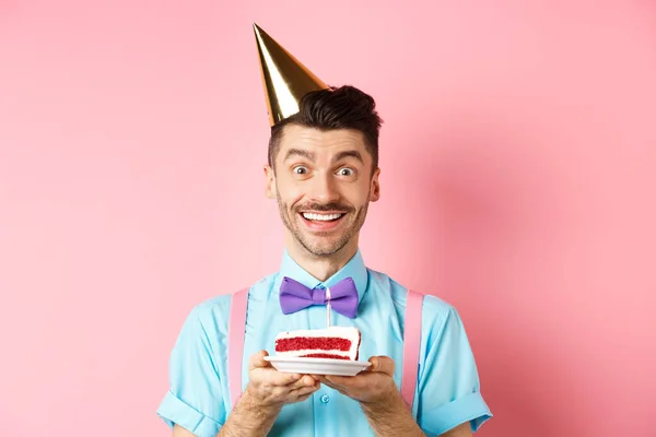 Holidays and celebration concept. Cheerful young man celebrating birthday in party hat, holding b-day cake with candle and making wish, smiling happy at camera, pink background