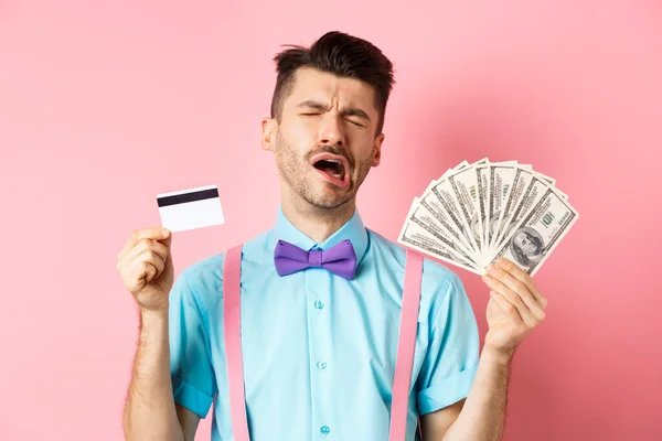 Triste hombre llorando buscando miserable, mostrando tarjeta de crédito de plástico y dinero, de pie molesto sobre fondo rosa — Foto de Stock