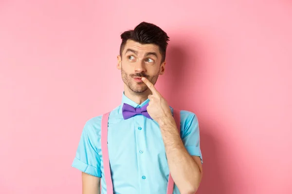 Imagem de homem engraçado bonito em arco-gravata olhando pensativo lábio esquerdo e comovente, pensando o que escolher, tomar uma decisão, de pé sobre fundo rosa — Fotografia de Stock