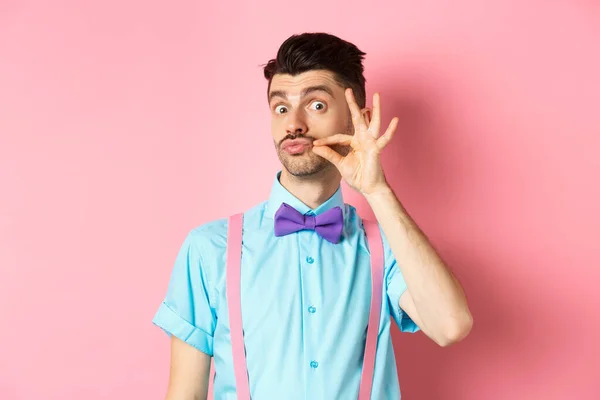 Engraçado jovem tocando seu bigode francês e lábios pucker, olhando tolo para a câmera, de pé em arco-gravata e suspensórios em fundo rosa — Fotografia de Stock