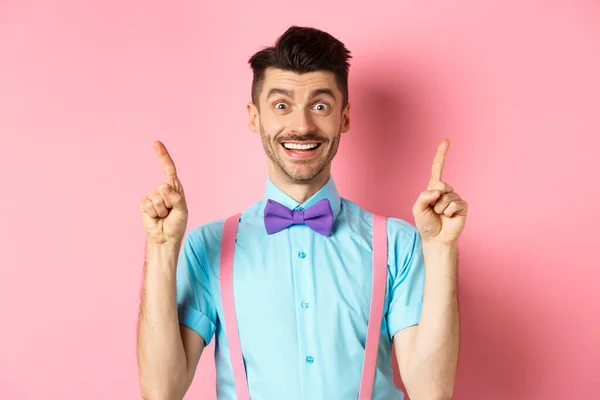 Retrato de un joven alegre con bigote y cerdas apuntando con los dedos hacia arriba, mostrando el logo superior, sonriendo feliz ante la cámara, de pie sobre fondo rosa — Foto de Stock