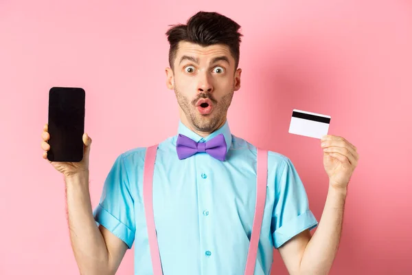 Concepto de comercio electrónico y compras. Hombre sorprendido mostrando pantalla de teléfono inteligente en blanco con tarjeta de crédito de plástico, de pie sorprendido sobre fondo rosa — Foto de Stock