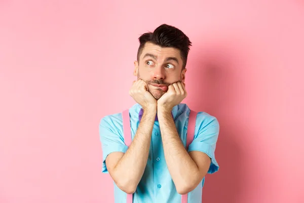 Imagen de joven tonto y lindo en tirantes, soñando con algo, mirando a la izquierda con cara curiosa, de pie sobre fondo rosa —  Fotos de Stock