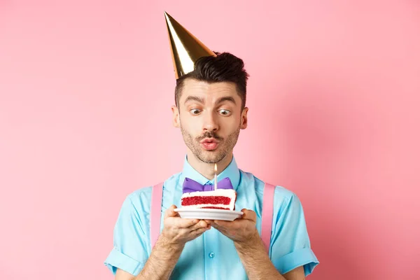 Vacaciones y concepto de celebración. Feliz joven en la fiesta de cumpleaños sombrero soplando vela en la torta, pidiendo deseo en b-day, de pie sobre fondo rosa — Foto de Stock