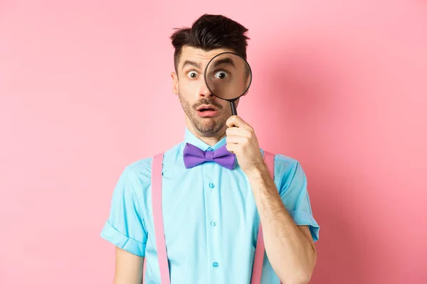Funny guy look through magnifying glass with surprised face, seeing something interesting, standing on pink background