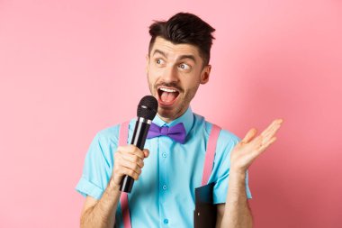 Erkek sunucunun konuşma yaparken görüntüsü, koltuk altında pano olan mikrofon ile konuşması, insanları şenlikli bir olaya dahil etmesi, pembe arka planda dikilmesi.