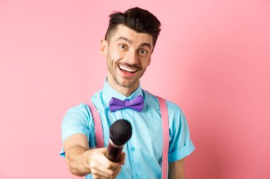 Bıyıklı ve papyonlu gülümseyen adam yorum ya da röportaj istiyor mikrofonla elini uzatıp pembe arka planda duruyor.