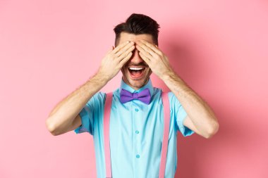 Neşeli genç adam, sürpriz bekliyor, gözleri kapalı ve mutlu bir gülümseme, pembe arka planda neşeli bir şekilde duruyor.