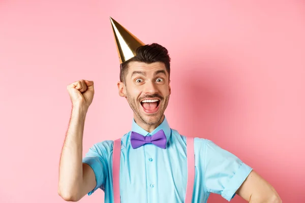 Holidays and celebration concept. Happy birthday boy with moustache and bow-tie celebrating event in party cone hat and festive clothes, raising hand up and shout yes