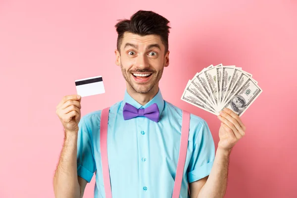 Alegre hombre caucásico con bigote y pajarita mostrando tarjeta de crédito de plástico con dinero en dólares, sonriendo a la cámara, de pie sobre fondo rosa — Foto de Stock