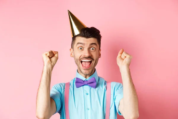 Férias e conceito de celebração. Jovem alegre comemorando festa de aniversário em chapéu de cone e gravata borboleta, gritando sim com emoção e fazendo gesto de bomba de punho, fundo rosa — Fotografia de Stock