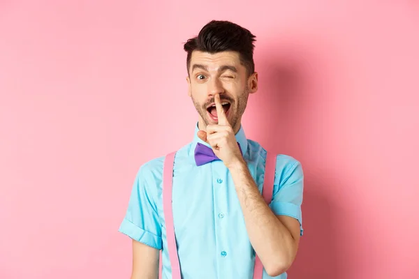 Funny young man planning a surprise, hushing and winking at camera, asking to keep quiet, telling a secret, standing on pink background — Stock Photo, Image
