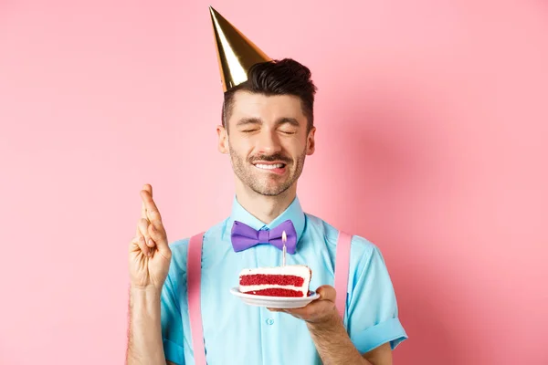 Vakantie en feest concept. Hoopvolle verjaardag man in feesthoed maken wens, het houden van taart met kaars en kruis vingers voor droom uitkomen, roze achtergrond — Stockfoto