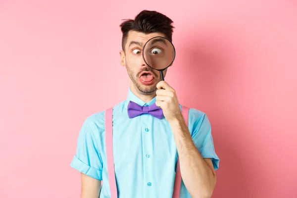 Rolig man i fluga titta genom förstoringsglas, kisa och göra dumma ansikten, står på rosa bakgrund — Stockfoto