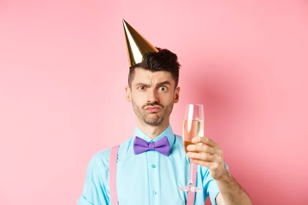 Holidays and celebration concept. Troubled young man in party hat, frowning with doubtful face, raising glass of champagne perplexed, standing on pink background