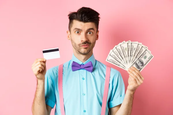 Hombre guapo en pajarita mostrando tarjeta de crédito de plástico y dinero en dólares, haciendo una oferta, de pie sobre fondo rosa — Foto de Stock