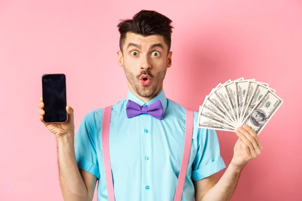 Concepto de comercio electrónico y compras. Hombre sorprendido mostrando pantalla de teléfono inteligente en blanco y dinero, diciendo wow con la cara sorprendida, echa un vistazo a la oferta en línea, fondo rosa —  Fotos de Stock