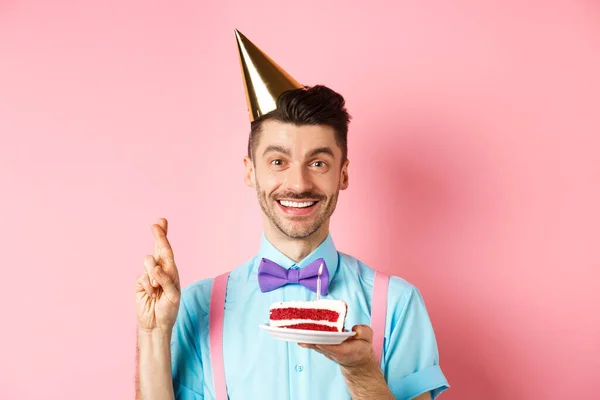 Vakantie en feest concept. Gelukkige jongeman genieten van verjaardagsfeestje, het dragen van kegel hoed en kruisvingers, het maken van wens op bday cake met kaars, roze achtergrond — Stockfoto