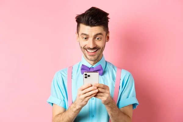 Online nakupování. Šťastný chlap při pohledu na displej chytrého telefonu, čtení zprávy a usmívání, stojí na růžovém pozadí — Stock fotografie