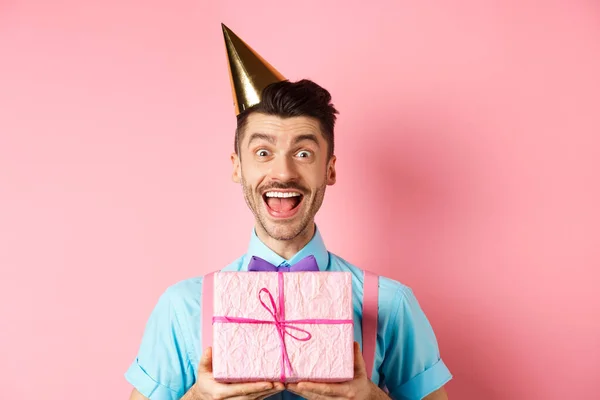 Vacaciones y concepto de celebración. Primer plano de chico feliz en sombrero de fiesta recibir regalo de cumpleaños, grito de alegría y alegría, de pie sobre fondo rosa — Foto de Stock