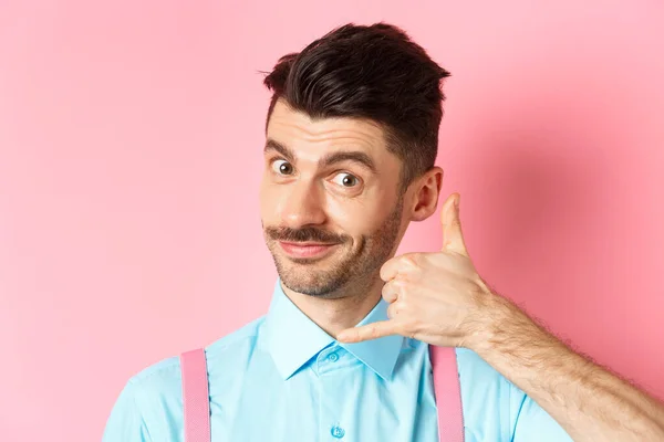 Nær venn av en kjekk, ung fyr med bart, som viser en telefongest, ber om å få ringe ham, står på rosa bakgrunn – stockfoto