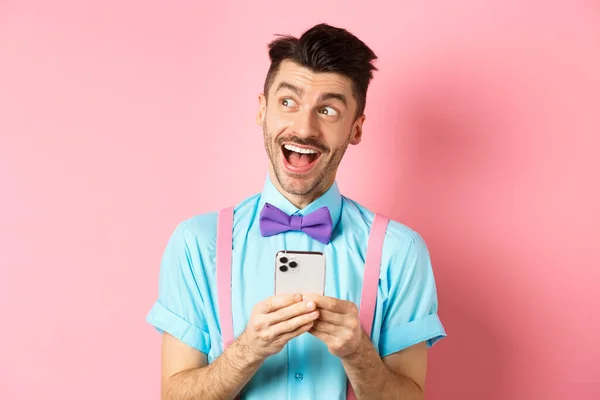 テクノロジーの概念。夢のような顔を脇に見て興奮した男,スマートフォンでオンライン賞を受賞,ピンクの背景の上に陽気に立って — ストック写真