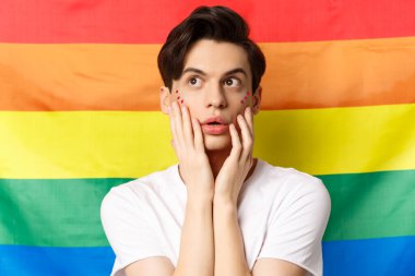 Yüzünde parıltı makyajı olan, sol üst köşeye bakan, heyecanlı, lgbtq gökkuşağı bayrağına karşı duran şehvetli ve güzel bir gay portresi.