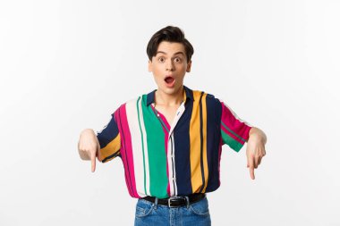 Şaşırmış ve şaşırmış bir adamın logosunu gösteren, parmaklarını aşağıya gösteren ve kameraya heyecanla bakan beyaz arka planda duran görüntüsü.