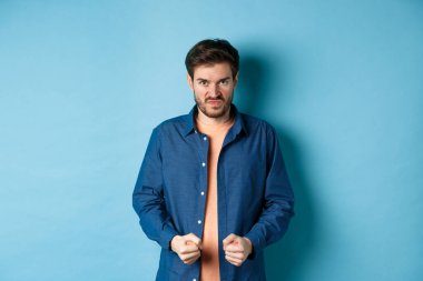 Angry and pissed-off man clench fists and stare with disdain at camera, trying to keep himself together, look with rage, standing on blue background