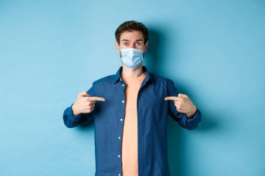 Covid-19 ve pandemik konsept. Sağlık maskeli sağlıklı genç adam mavi arka planda duran orta logoyu işaret ediyor.